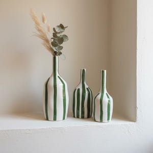 Op de afbeelding: Drie keramische vazen met een witte basis en verticale donkergroene strepen. De hoogste vaas aan de linkerkant bevat gedroogd pampasgras en eucalyptusstengels. Elke vaas heeft een smalle hals en een afgerond lichaam met een gladde afwerking.