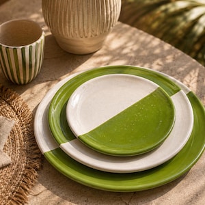 Op de afbeelding: Een set keramisch servies bestaande uit een stapel van twee ronde borden en een bijpassende beker. Elk item is afgewerkt met een tweekleurig ontwerp, waarbij een glanzend grasgroen glazuur wordt gecombineerd met een gespikkeld gebroken wit oppervlak.
