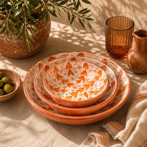 Pode incluir: Um conjunto de três taças de cerâmica empilháveis com um padrão pintado à mão de folhas e bagas cor de laranja sobre um fundo creme. As taças têm uma forma arredondada e uma estética quente e terrosa, empilhadas sobre uma superfície de tecido.