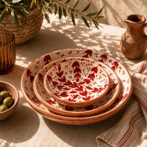 May include: A set of three nested ceramic bowls featuring a cream-coloured base with hand-painted red leaf patterns and small dots. The bowls have a rounded shape and a smooth finish, showcasing a consistent botanical design across all three sizes.