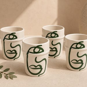 Op de afbeelding: Een set van vier gebroken witte keramische bekers, elk voorzien van een doorlopende donkergroene lijntekening van een menselijk gezicht. De bekers hebben een cilindrische vorm met een subtiele horizontale rand nabij de bovenste rand.