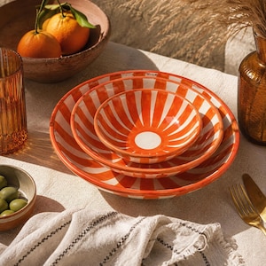 May include: A set of three nested ceramic bowls featuring a vibrant orange and white sunburst or striped pattern. Each bowl has a solid white circular centre surrounded by radiating orange and white stripes that extend to the outer rim.