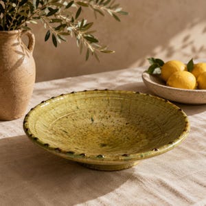 Peut inclure: Un bol en céramique peu profond et circulaire avec un vernis moucheté vert olive et jaune. Le bord présente une série de petites bosses surélevées régulièrement espacées. Le bol a une base large et plate avec une finition lisse et brillante.