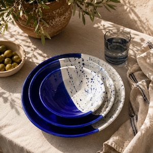 Peut inclure: Un ensemble de trois bols en céramique emboîtables au design bicolore. Chaque bol est divisé en une section bleu profond unie et une section blanche avec un motif bleu moucheté. Les bols sont empilés, affichant un style cohérent.