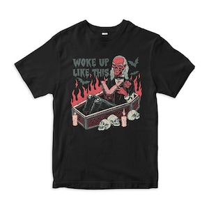 Puede incluir: Una camiseta negra con un gráfico de un vampiro sentado en un ataúd leyendo un libro, rodeado de llamas, murciélagos, velas y calaveras. El texto WOKE UP LIKE THIS está impreso en rojo y blanco sobre la figura del vampiro.