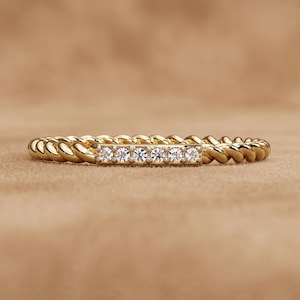 Könnte beinhalten: Ein goldener Ring mit einem gedrehten, seilartigen Band und einer flachen, rechteckigen Fassung in der Mitte. Diese Fassung ist mit sechs kleinen, klaren, rund geschliffenen Steinen besetzt, die in einer horizontalen Reihe angeordnet sind.