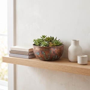 May include: A hammered copper bowl with a green patina finish containing a variety of green succulents. The bowl sits on a light wood shelf next to a stack of three books, a matte white ceramic vase, and a small white candle in a ceramic holder.