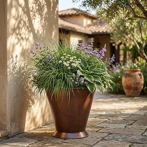 Puede incluir: Una maceta de cobre grande y cónica con un acabado texturizado y martillado. La maceta está llena de una variedad de follaje verde, que incluye hierbas altas, hostas de hojas anchas, pequeñas flores blancas y espigas de flores moradas.