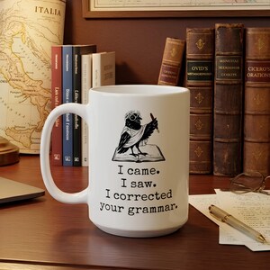 May include: A white ceramic mug featuring a black graphic of a bird wearing a laurel wreath, standing on an open book while holding a quill. Below the graphic, the text reads: I came. I saw. I corrected your grammar. The mug has a handle on the left.