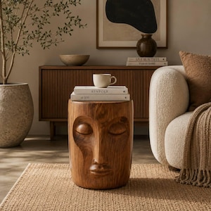 May include: A cylindrical wooden stool featuring a carved human face with closed eyes, a nose, and lips on the front surface. Two white books titled Kinfolk and at 100 are stacked on top of the stool, topped with a plain light-coloured ceramic mug.