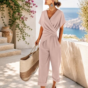 Könnte beinhalten: Ein hellrosa Jumpsuit mit einem Wickeloberteil, V-Ausschnitt, kurzen Ärmeln und einer taillierten Passform. Die Hose hat einen geradlinigen Schnitt mit umgeschlagenen Säumen. Eine große, geflochtene Strohtasche mit geschichtetem Design wird seitlich getragen.
