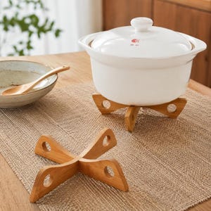 May include: A white ceramic cooking pot with a lid featuring the red text MK 47, resting on a light brown wooden cross-shaped trivet. A second identical wooden trivet sits in the foreground, and a ceramic bowl with a wooden spoon is to the left.