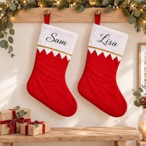 May include: Two red and white Christmas stockings featuring a white cuff with a gold trim line. The left stocking is personalised with the name Sam in black script, and the right stocking is personalised with the name Lisa in black script.