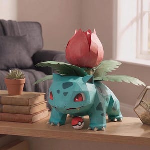 Puede incluir: Un modelo de papel de la criatura Venusaur, con un cuerpo verde azulado con manchas oscuras, ojos rojos y un gran bulbo floral rosa en la espalda. Un pequeño juguete esférico rojo y blanco descansa entre sus patas delanteras sobre una superficie de madera.
