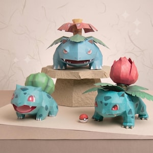 May include: Three papercraft figures of blue creatures with green bulbous or floral features on their backs. One figure stands on a stone-like pedestal, while two others sit below. A small red and white spherical object rests on the surface.