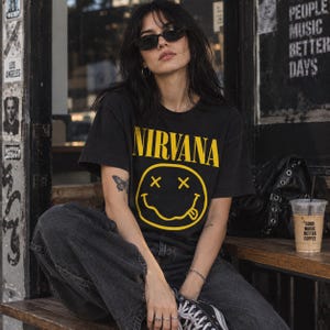 May include: A black short-sleeved t-shirt featuring the yellow text NIRVANA above a yellow graphic of a smiley face with X-shaped eyes and a tongue sticking out. The shirt is worn with dark grey denim jeans and black and white canvas trainers.