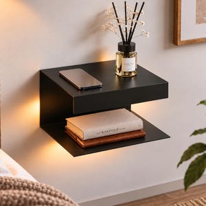 Può includere: Una mensola a muro flottante nero opaco a due livelli con illuminazione LED calda integrata. La superficie superiore ospita un diffusore di fragranze in vetro e uno smartphone, mentre il ripiano inferiore sostiene due libri impilati, tra cui The Kinfolk Home.