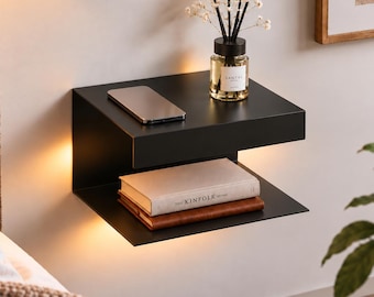 Op de afbeelding: Een matzwarte, aan de muur gemonteerde zwevende plank met twee niveaus en geïntegreerde warme LED-verlichting. Het bovenoppervlak bevat een glazen geurverspreider en een smartphone, terwijl de onderste plank twee gestapelde boeken bevat, waaronder The Kinfolk Home.