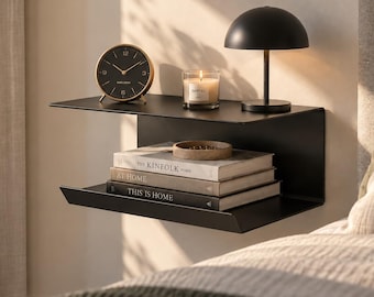Op de afbeelding: Een matzwarte metalen wandplank met twee niveaus. Het bovenste oppervlak bevat een ronde zwarte analoge klok, een glazen kaars en een zwarte koepellamp. De onderste plank bevat een stapel van drie boeken met de titels The Kinfolk Home, At Home en This Is Home.