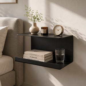 Può includere: Una mensola da parete sospesa in metallo nero opaco con due ripiani orizzontali. La superficie superiore ospita un vaso in ceramica beige con vegetazione, una candela in vetro marrone e un orologio analogico rotondo. Il ripiano inferiore contiene tre libri.
