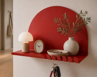 Op de afbeelding: Een rood wandrek met een halfrond achterpaneel en een plat oppervlak met geïntegreerde haken. Op het rek staan een kleine witte paddenstoelvormige lamp, een ovale houten klok, een beige kom en een vaas met groene bladeren en takken.