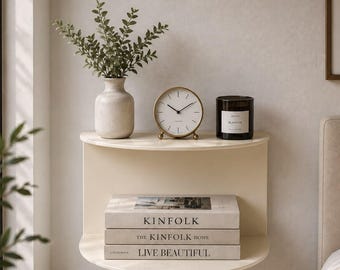 May include: A semi-circular wall-mounted shelf in a cream finish. The top surface holds a beige ceramic vase with green foliage, a round gold-rimmed analog clock with a white face, and a black candle jar. Three stacked books sit on the lower shelf.