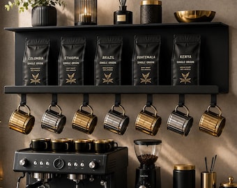 Op de afbeelding: Een zwarte wandplank met vijf zwarte koffiezakken met de labels Colombia, Ethiopia, Brazil, Guatemala en Kenya. Zeven goudkleurige en zwarte mokken hangen aan haken eronder. Een zwarte espressomachine en koffiemolen staan op het aanrecht.