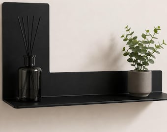 Op de afbeelding: Een matzwarte metalen wandplank met een L-vormig achterpaneel. Aan de linkerkant staat een donkere glazen geurstokjeshouder met zwarte stokjes. Aan de rechterkant staat een kleine grijze keramische pot met een groene plant.