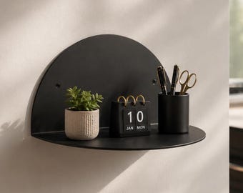Op de afbeelding: Een halfronde matzwarte wandplank met een plat oppervlak en een gebogen achterpaneel. Op de plank staat een kleine, getextureerde witte pot met een groene vetplant, een zwarte flipkalender met 10 JAN MON en een zwarte pennenhouder.