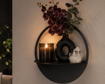 Op de afbeelding: Een cirkelvormige wandplank van zwart metaal met een plat horizontaal oppervlak. Op de plank staat een donkere glazen kaarshouder met een brandende kaars, een zwarte donutvormige vaas met donkerpaarse bloemen en een helder kwartskristal.
