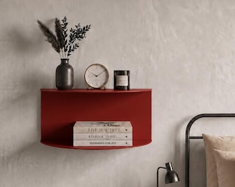Op de afbeelding: Een halfronde wandplank in een dieprode afwerking. Het bovenoppervlak bevat een donkere vaas met gedroogd loof, een ronde analoge klok en een zwarte kaars. Een stapel van drie boeken met de titels Live Beautiful, Kinfolk Travel en Designing for Joy.