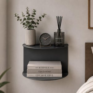 May include: A dark gray, semi-circular wall-mounted shelf holding a ribbed vase with greenery, a round analog clock, and a glass reed diffuser. Below, three stacked books titled The Kinfolk Home, This Is Home, and Live Beautiful are displayed.