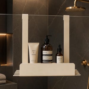 May include: A cream-colored metal shower caddy with two vertical hooks for hanging over a glass partition. The shelf holds a tube labeled Salt & Stone, a brown glass bottle labeled Aesop, and a small dropper bottle labeled Meraki.