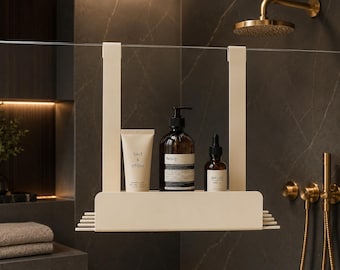 Op de afbeelding: Een crèmekleurig metalen doucheplankje met twee verticale haken om over een glazen wand te hangen. Het plankje bevat een tube met het label Salt & Stone, een bruine glazen fles met het label Aesop en een klein Meraki-druppelflesje.