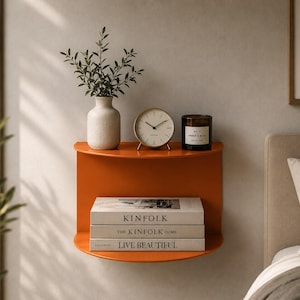 May include: A semi-circular orange wall-mounted shelf holding a white vase with greenery, a round analog clock, and a dark candle. The lower shelf displays three stacked books titled Kinfolk, The Kinfolk Home, and Live Beautiful.