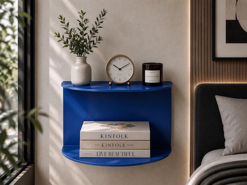 May include: A semi-circular blue wall-mounted shelf featuring two tiers. The top tier holds a white vase with greenery, a round gold-rimmed analog clock, and a dark glass candle. The bottom tier holds three stacked books titled KINFOLK, THE KINFOLK HOME, and LIVE BEAUTIFUL.