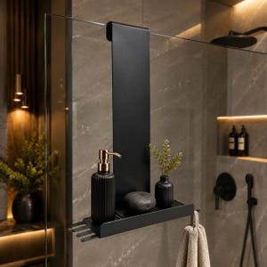 May include: A matte black over-the-glass shower caddy featuring a vertical mounting panel, a horizontal storage shelf, and integrated hooks. The shelf holds a ribbed black soap dispenser with a gold pump, a small black vase with greenery, and a dark stone.