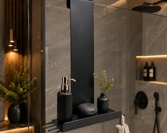 Op de afbeelding: Een matzwarte douche-organizer voor over de glaswand met een verticaal paneel, een horizontale plank en haken. Op de plank staan een geribbelde zwarte zeepdispenser met gouden pomp, een kleine zwarte vaas met groen en een donkere steen.