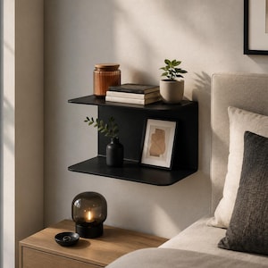 May include: A matte black metal wall-mounted shelf featuring two horizontal surfaces connected by a vertical back panel. The shelf holds a glass jar, a stack of books, a small potted plant, a black vase with greenery, and a framed abstract print.