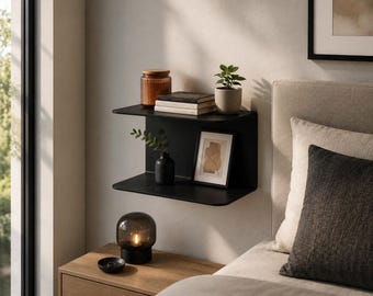 Op de afbeelding: Een matzwarte metalen wandplank met twee horizontale oppervlakken verbonden door een verticaal achterpaneel. De plank bevat een glazen pot, een stapel boeken, een kleine potplant, een zwarte vaas met groen en een ingelijste abstracte print.