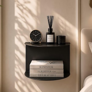 May include: A matte black semi-circular wall-mounted shelf holding a black analog clock, a reed diffuser, a small black candle, and three stacked books titled Kinfolk, The Kinfolk Home, and Live Beautiful, positioned against a neutral wall.