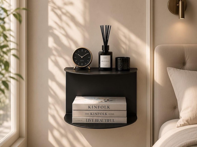 May include: A matte black semi-circular wall-mounted shelf holding a black analog clock, a reed diffuser, a small black candle, and three stacked books titled Kinfolk, The Kinfolk Home, and Live Beautiful, positioned against a neutral wall.