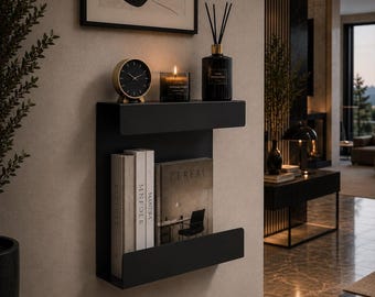 Op de afbeelding: Een matzwarte metalen wandplank met twee horizontale niveaus. Het bovenste niveau bevat een ronde analoge klok, een brandende kaars in een glazen pot en een zwarte geurstokjeshouder. Het onderste niveau bevat drie boeken, waaronder een met de titel Cereal.