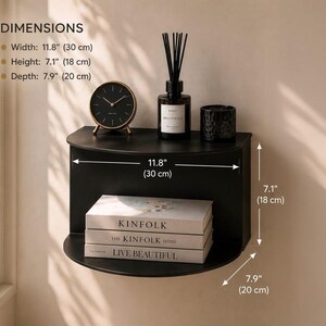 May include: A black semi-circular wall shelf with a flat top and curved front. Dimensions are 11.8 inches wide, 7.1 inches high, and 7.9 inches deep. The shelf holds a round clock, a reed diffuser, a candle, and three stacked books.