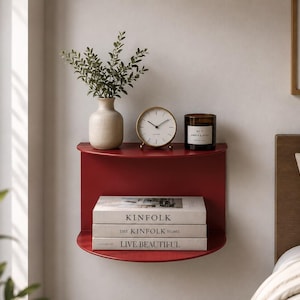 May include: A semi-circular wall-mounted shelf in a deep red finish. The top surface holds a beige ceramic vase with green foliage, a round gold-rimmed analog clock, and a dark glass candle. Three stacked books titled KINFOLK, THE KINFOLK HOME, and LIVE BEAUTIFUL sit on the lower shelf.