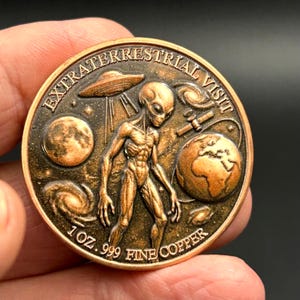 May include: A round copper coin featuring an alien figure in the center, a flying saucer, a planet, a galaxy, and a satellite. The text EXTRATERRESTRIAL VISIT is at the top, and 1 OZ .999 FINE COPPER is inscribed at the bottom of the coin.