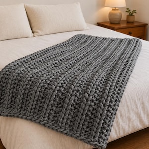 May include: A dark grey chunky knit throw blanket featuring a prominent vertical ribbed texture. The blanket is crafted with thick yarn, creating a dense, tactile pattern of raised columns and recessed stitches across the entire rectangular surface.