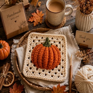Puede incluir: Un cuadrado de ganchillo de color crema que presenta una calabaza naranja tridimensional con un tallo verde centrado en la tela. La pieza se muestra en una bandeja tejida junto a un ganchillo de madera, un ovillo de lana y una caja de regalo.