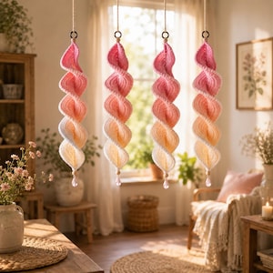 May include: Four hanging spiral decorations made of crocheted yarn in a vertical ombre gradient, transitioning from deep pink at the top to light peach and white at the bottom. Each piece is suspended by a cord and features a clear crystal bead.