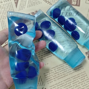 May include: Three clear, flexible plastic tubes filled with a light blue liquid and several dark blue, rounded, pebble-like shapes suspended inside. Each tube features a ribbed, light blue screw-on cap at the bottom and a crimped top seal.